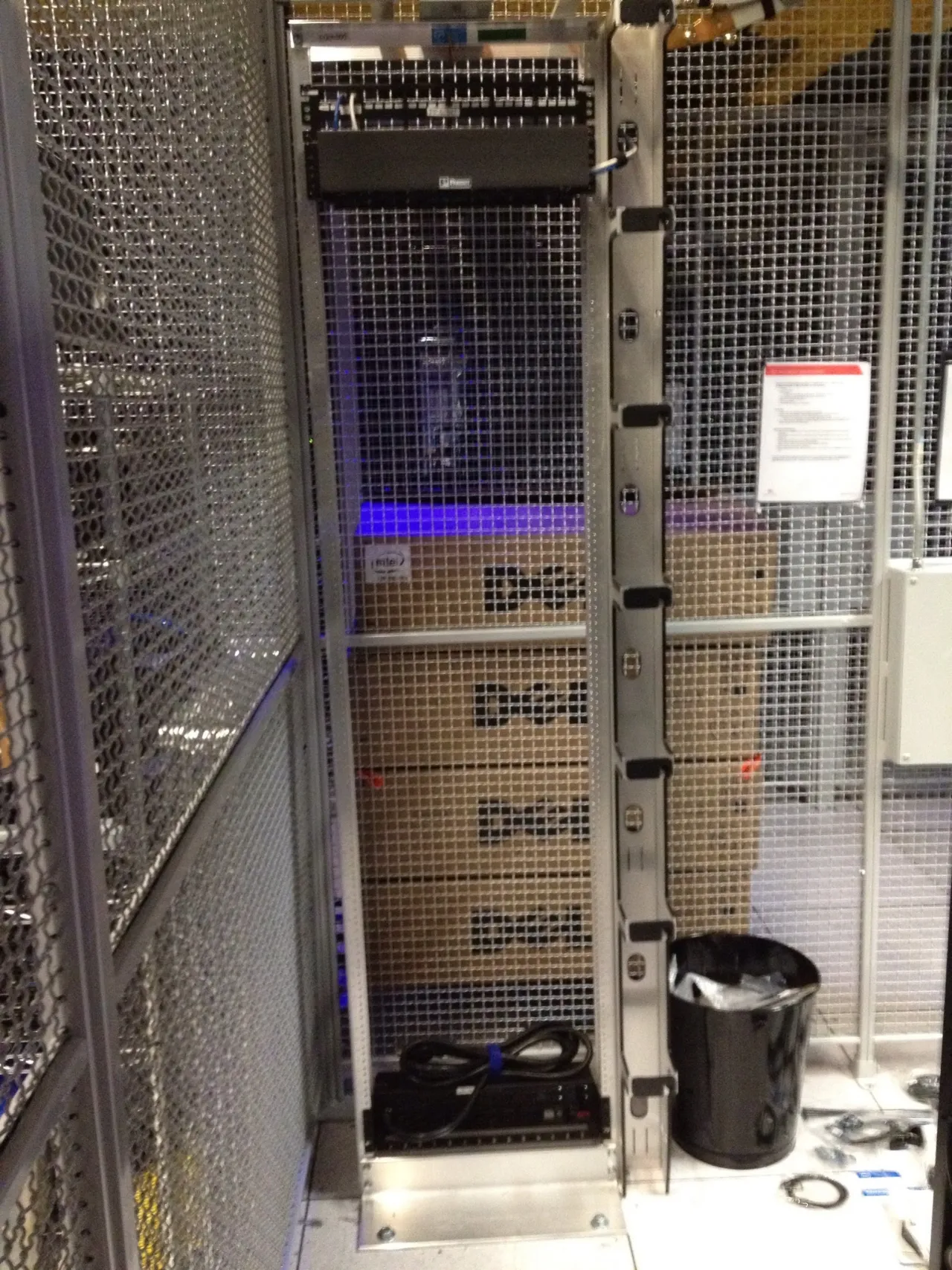 An empty rack in a wire-mesh data center cage, with boxed Dell servers stacked behind it and a power strip at its base